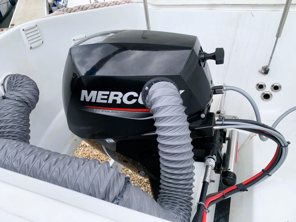 A black Mercury outboard motor is mounted on a boat, with gray hoses connected. The motor features a red and white logo. The background includes part of the boat interior with visible screws and a sandy surface.