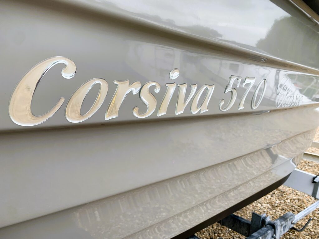 The side of a boat displayed with the name "Corsiva 570" in raised, polished letters. The boat's surface has a light beige color, and the background is blurred, showing a gravel surface where the boat is positioned.