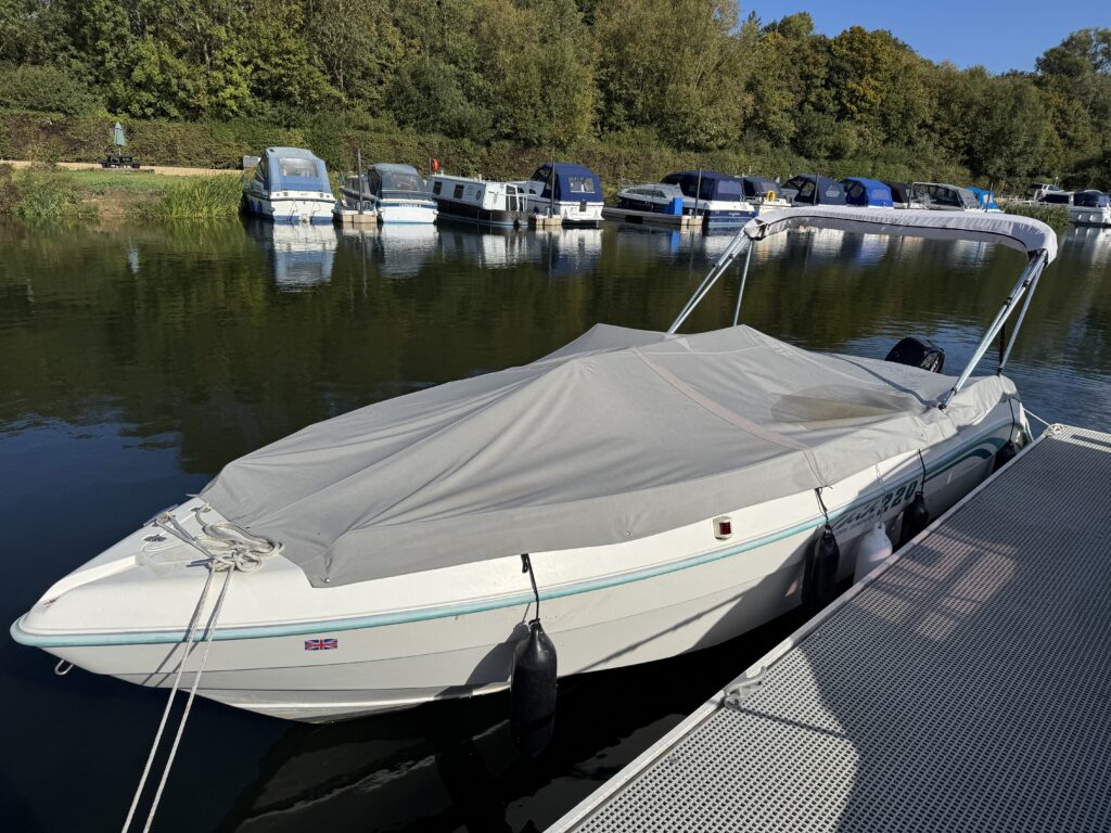 A white boat with a gray cover docked at a marina. The boat is positioned next to several other boats, reflecting the surrounding greenery and water in the calm scene.