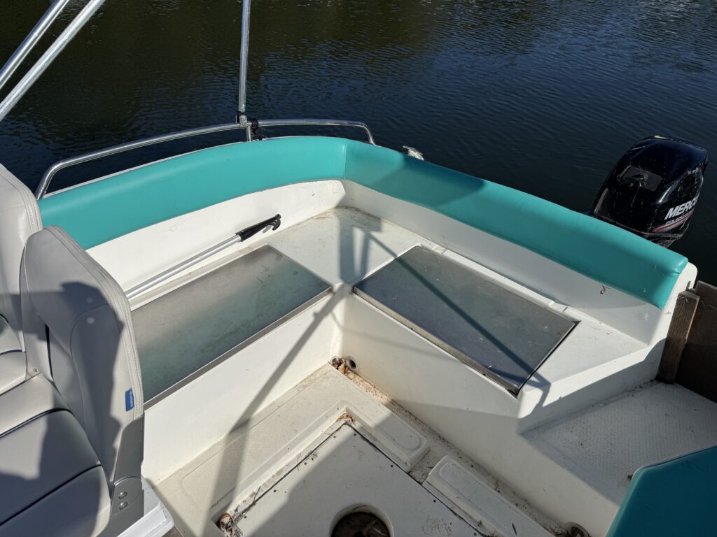 The interior of a small boat featuring a turquoise bench area with metal covers. The bench is equipped with open compartments and is situated near the boat's motor, which is visible in the background along with the water surface.