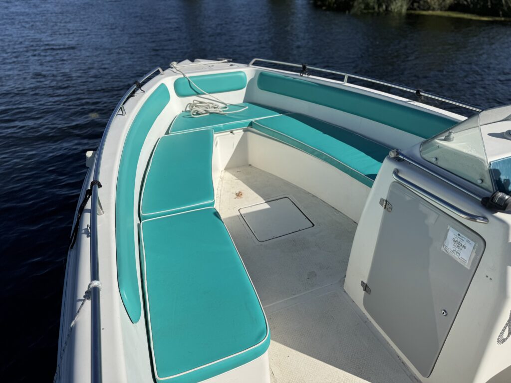 A boat interior featuring turquoise seating cushions arranged around the perimeter, with a small enclosed area in the front. The surface is light-colored, and the background shows dark water reflecting sunlight.