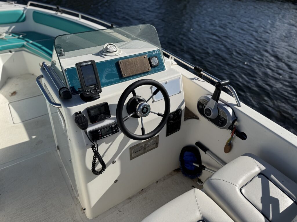 An overview of a boat’s console, featuring a steering wheel at the center, a digital display on the left side, and various controls on the right. The background shows water reflecting light, and seating is visible on the right side.