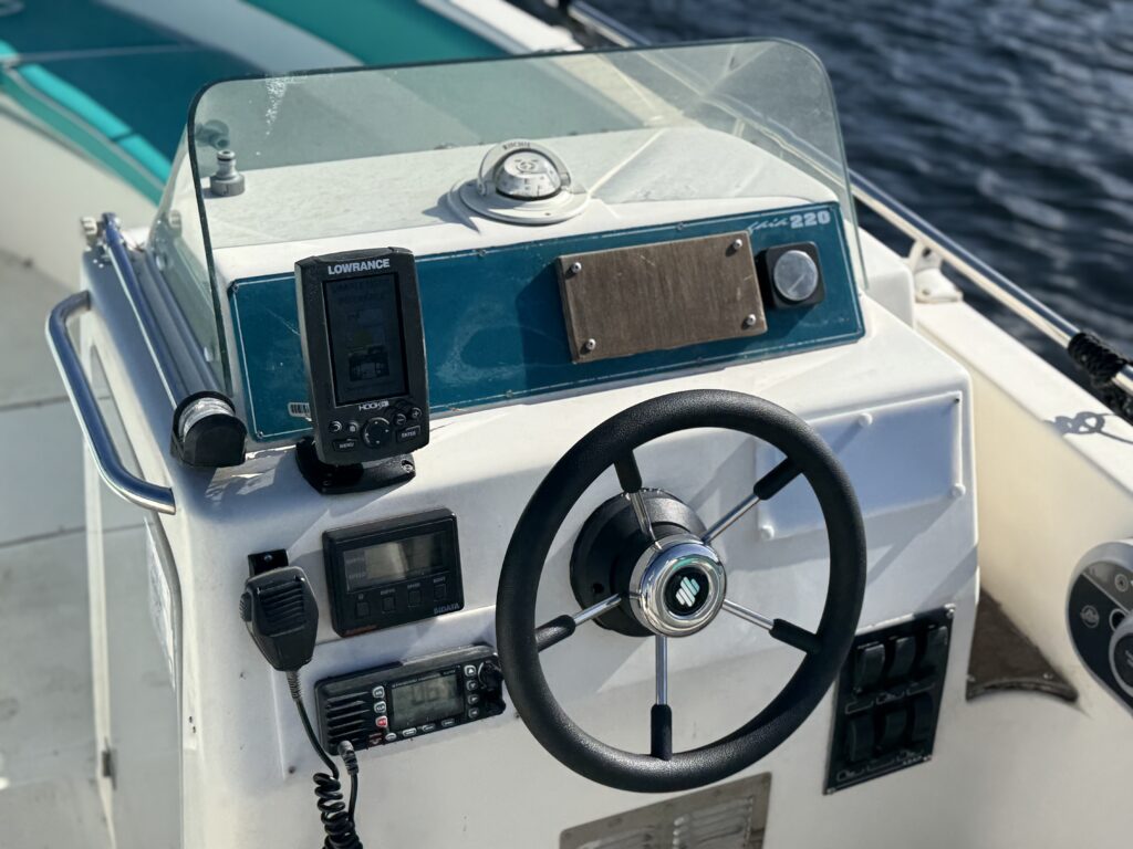 The image shows the steering console of a boat, featuring a black steering wheel, a Lowrance fishfinder, and various control buttons and gauges. The background includes part of the boat and water, indicating a boat interior.