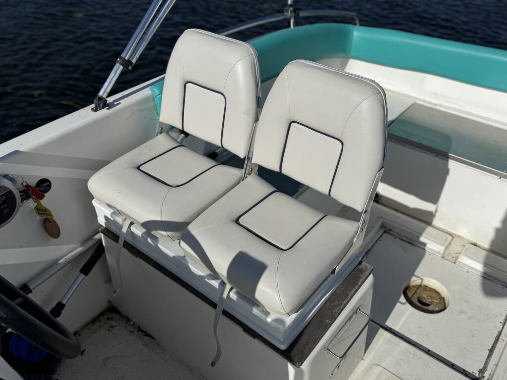 Two white marine-style boat seats with black piping, positioned on a flat surface in a boat. The background features teal accents and the water is visible, reflecting light.