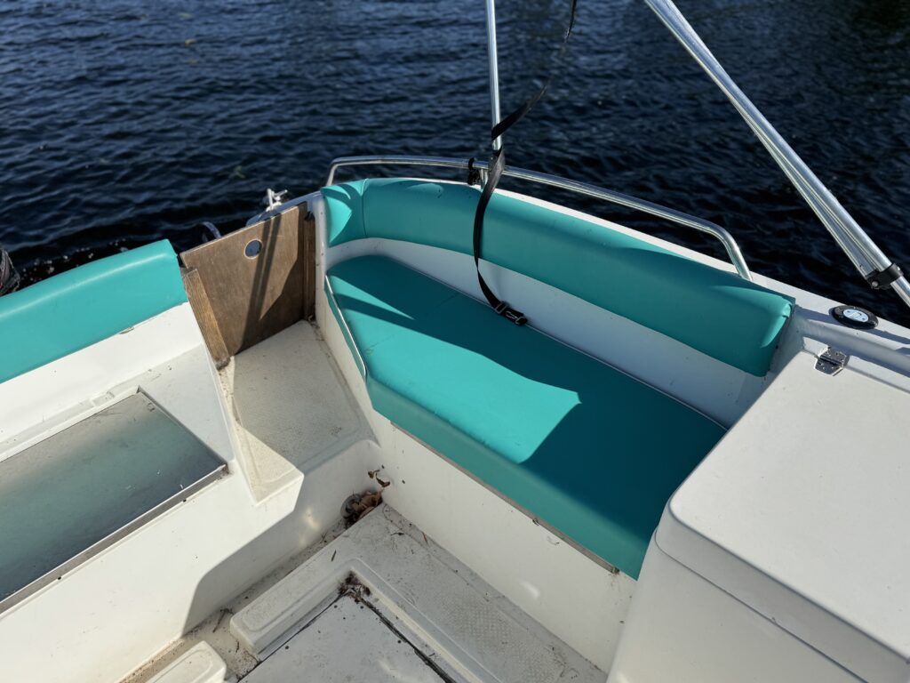 A close-up view of a boat interior featuring a turquoise cushioned seat along one side. The flooring is light-colored and there is a partial view of the boat's side panel and a metal railing. The background shows dark water.
