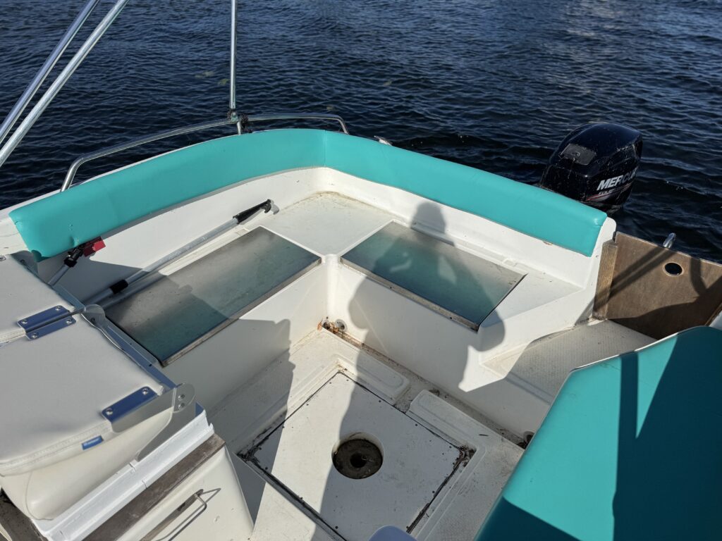 The interior of a boat showing a curved seating area with light turquoise upholstery, two clear hatches on the deck, and a shadow of a person. The background features dark water and a motor at the back of the boat.