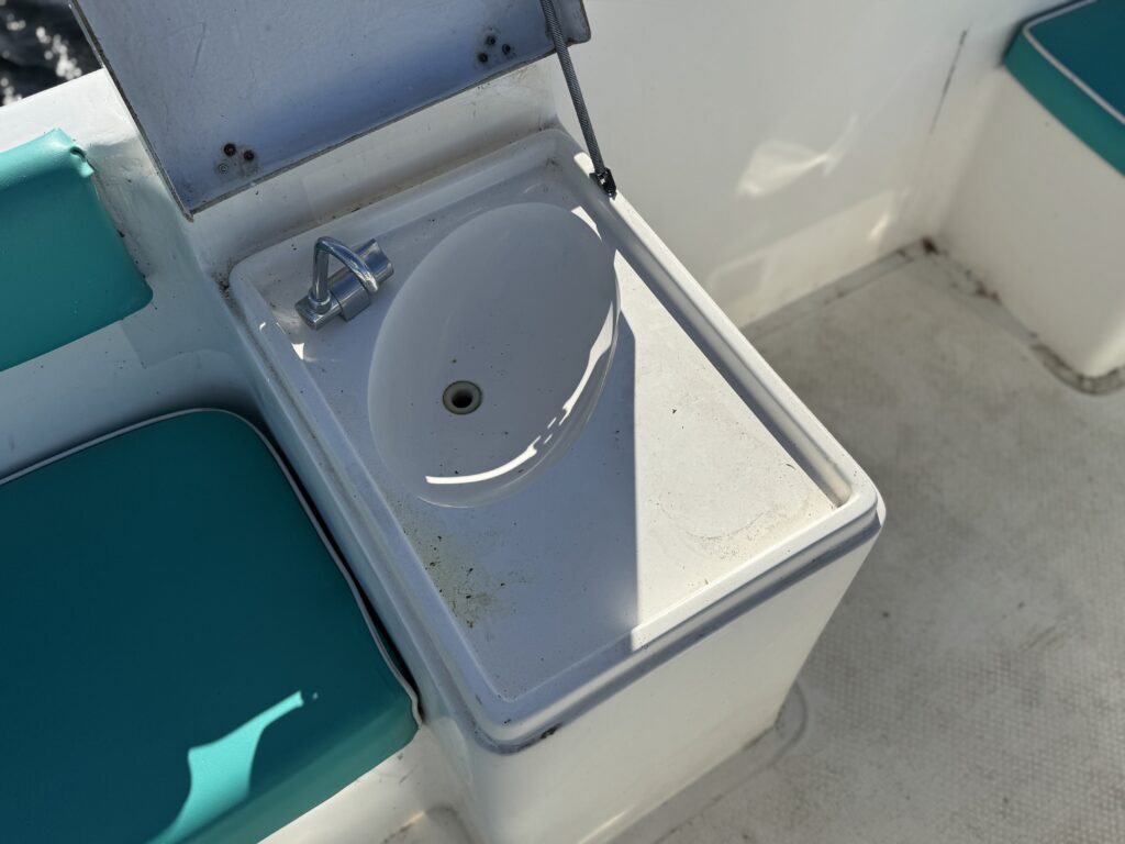 A small, white sink with a faucet is installed in a boat's cabin. The surrounding area features turquoise seating, and sunlight reflects off the sink's surface.