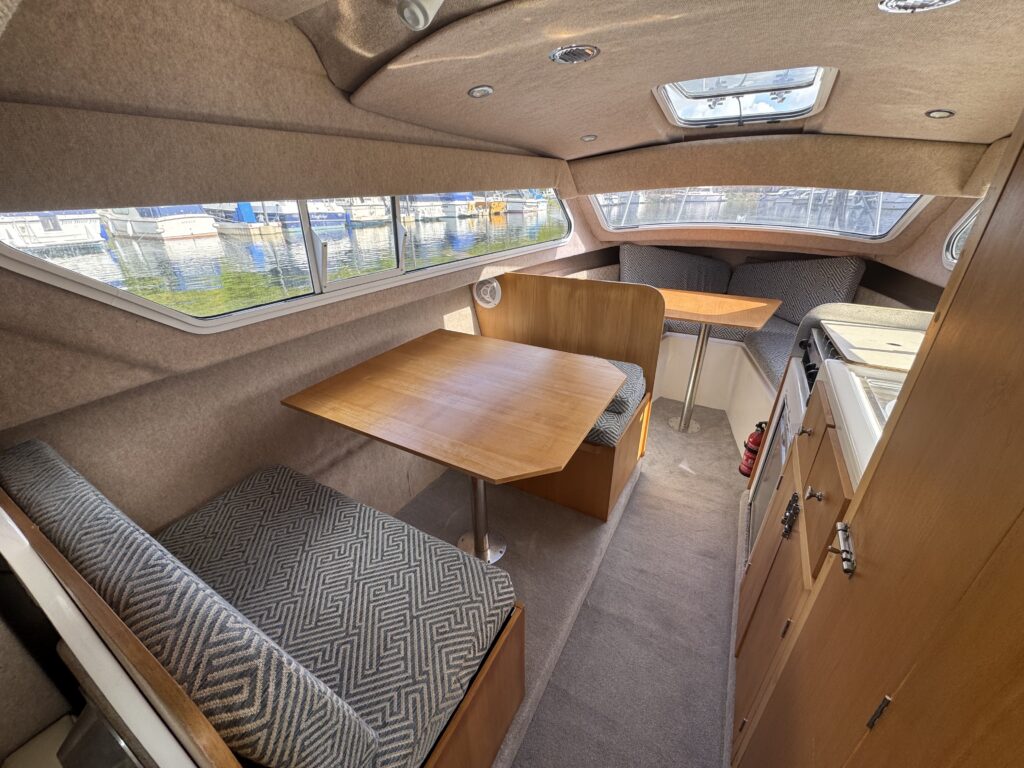 The interior of a boat featuring a cozy seating area with a patterned cushion and a wooden table. A compact kitchen counter is visible on the right, with storage cabinets below and large windows providing natural light.