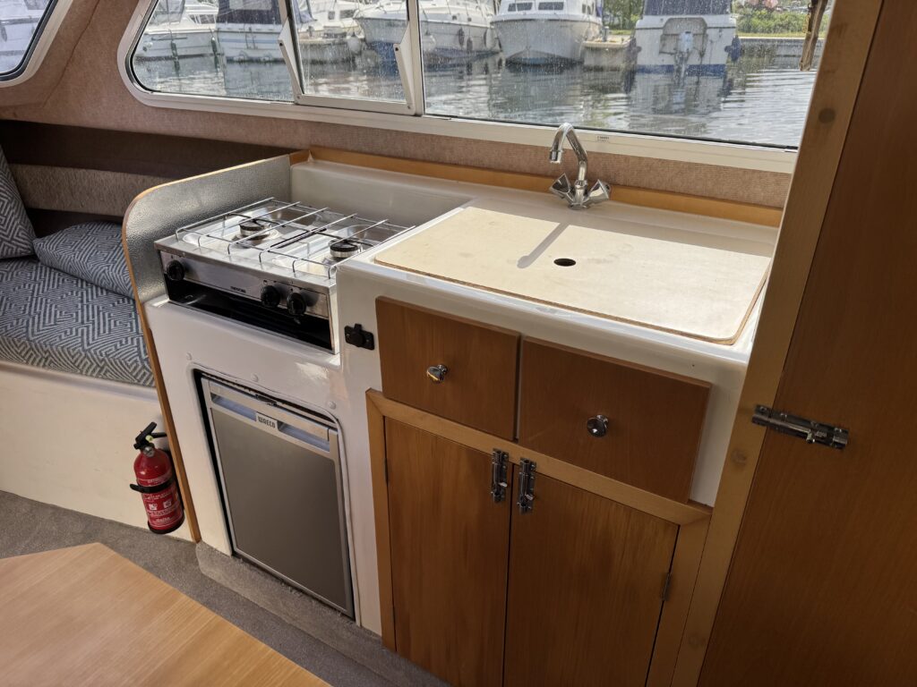 A compact galley kitchen on a boat featuring a gas stove, stainless steel sink, and a refrigerator beneath wooden cabinets. The setting includes a cozy seating area visible in the background, with windows showing boats moored outside.