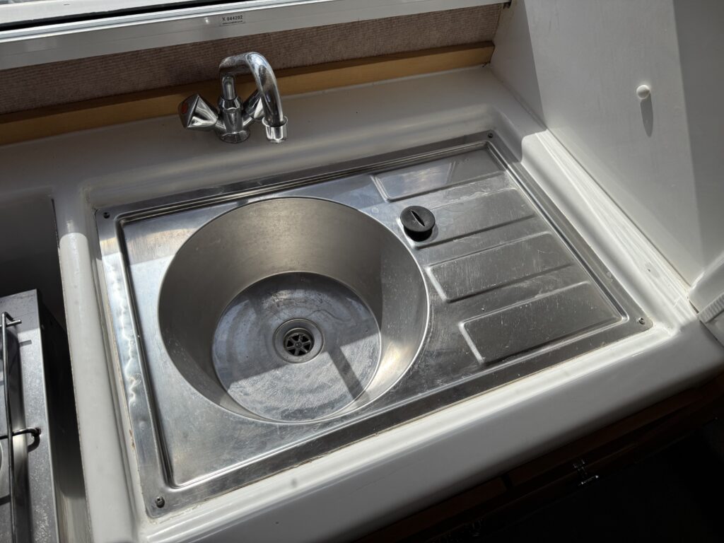 A stainless steel sink with a round basin and a flat surface is installed in a countertop. The faucet is attached above the sink, and there is a circular control knob near the basin. Natural light comes from a nearby window.