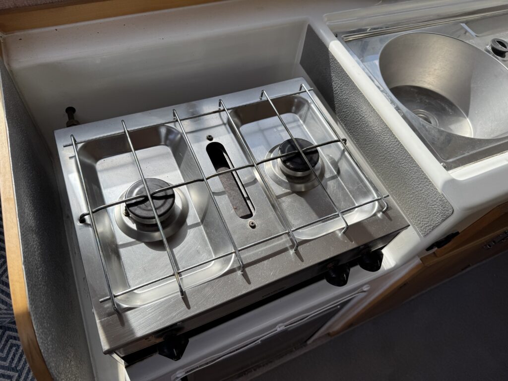 A stainless steel two-burner gas stove mounted on a wooden countertop. The stove features a protective grid over the burners and is located next to a round stainless steel sink.