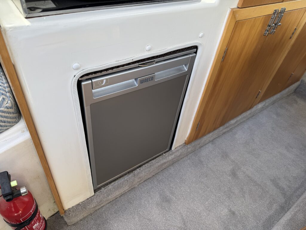 A compact stainless steel refrigerator is built into a wooden cabinet on a carpeted floor. The refrigerator's door features a handle, and the surrounding area includes a fire extinguisher and part of a textured wall.