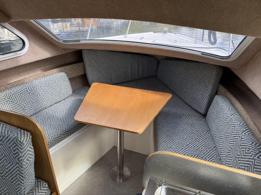 An interior shot of a boat featuring a cozy seating area with patterned cushions. A wooden table stands at the center, supported by a metal pedestal. The walls are lined with soft fabric, and large windows provide natural light.