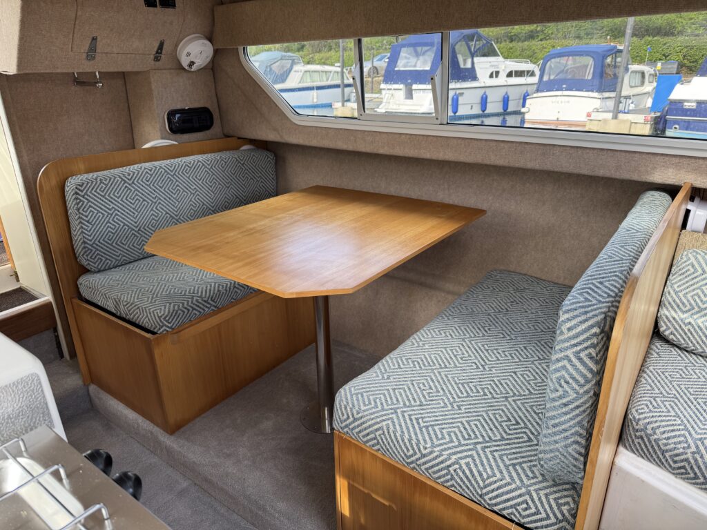 A compact boat interior featuring a wooden table and two upholstered benches with geometric pattern fabric. Natural light streams in through a window, providing a view of parked boats outside.