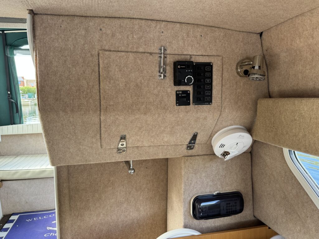 An interior view of a boat featuring a beige wall panel with a control panel and several switches. A round light fixture is mounted on the wall, while a small window shows part of the boat's exterior. The floor has a welcome mat.