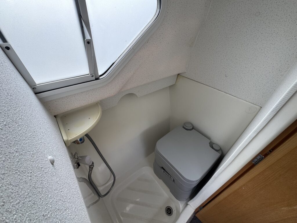 A compact bathroom space featuring a small toilet with a gray tank, a simple sink with a light-colored countertop, and a shower area with a molded floor. The walls are white with a small window allowing natural light in.