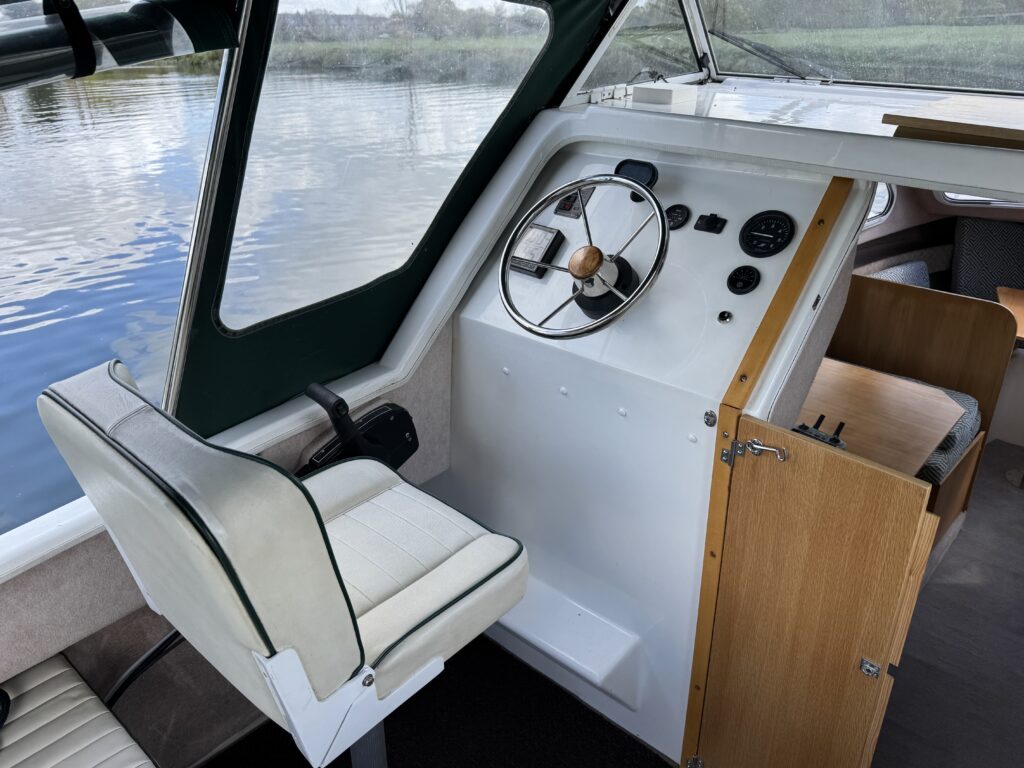 The interior of a boat featuring a white steering wheel, control panel with gauges, and a beige padded captain's seat. The background shows a body of water and a green landscape through a large window.