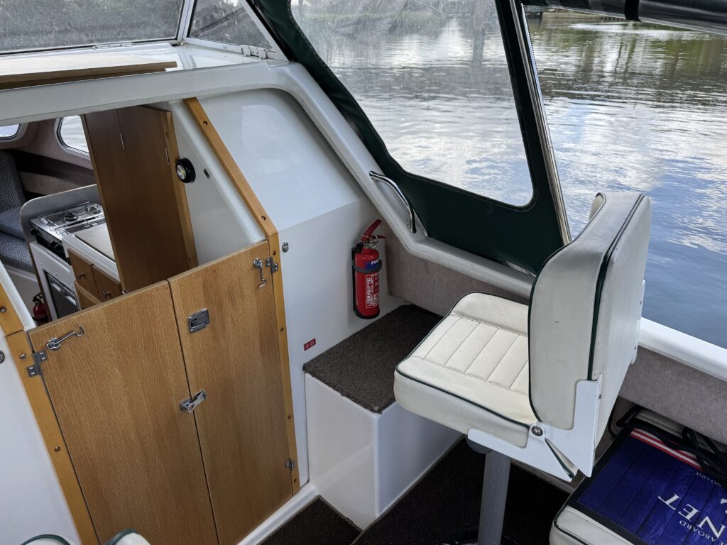 The interior of a boat showing a white leather captain's chair beside a wooden cabinet with a door. A red fire extinguisher is mounted on the wall, and the water is visible through the large window to the right.
