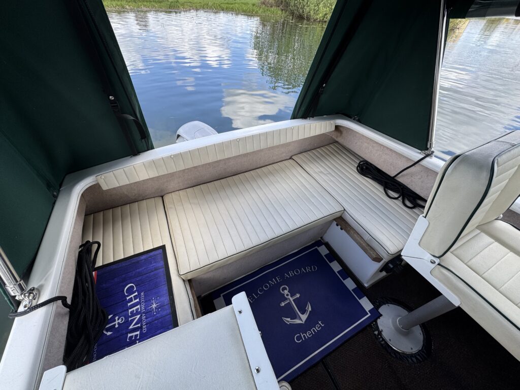 The interior of a boat showing a covered seating area with light-colored cushions and a dark blue welcome mat featuring an anchor design. The setting has green curtains and reflects water and greenery from outside.
