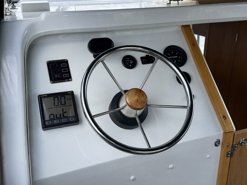 A close-up view of a boat's control panel featuring a wooden steering wheel at the center. To the left are digital displays and control buttons, and to the right are various gauges. The surface is white, with a wooden cabinet in the background.
