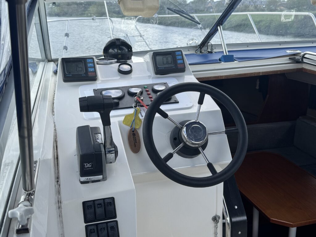 A close-up view of a boat's cockpit featuring a steering wheel, control panel with gauges, a throttle lever, and various buttons. In the background, the interior includes a wooden table and a window revealing water and greenery outside.