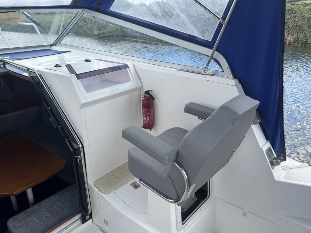 The interior of a boat, featuring a gray padded captain's chair with armrests positioned next to a dashboard. A fire extinguisher is mounted on the wall, and a large blue canopy covers the helm area, with a view of water visible through a window.