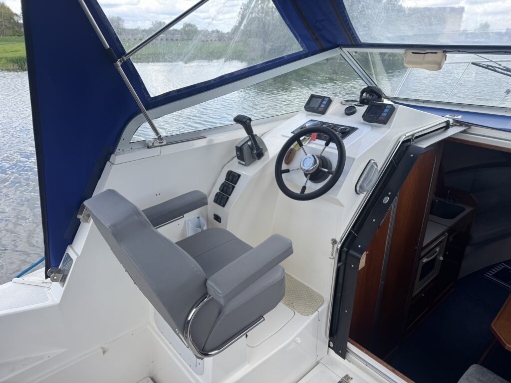 The interior of a boat cockpit features a gray captain's chair positioned beside a steering wheel and control panel with various gauges and controls. A blue canopy covers the area, and a body of water is visible through the open side.