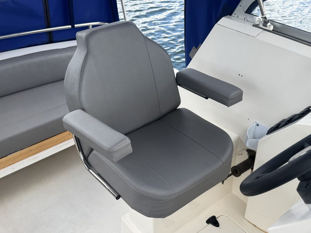 A gray padded captain's chair is positioned on a boat's helm area. The chair features armrests and a simple design, situated against a background of the boat’s interior and open water visible through the window.