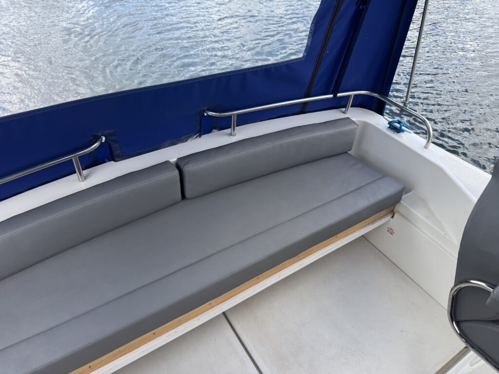 A gray cushioned bench seating area on a boat, with a blue canopy in the background. The bench is upholstered with a sleek fabric and is positioned against a light-colored deck, overlooking a body of water.