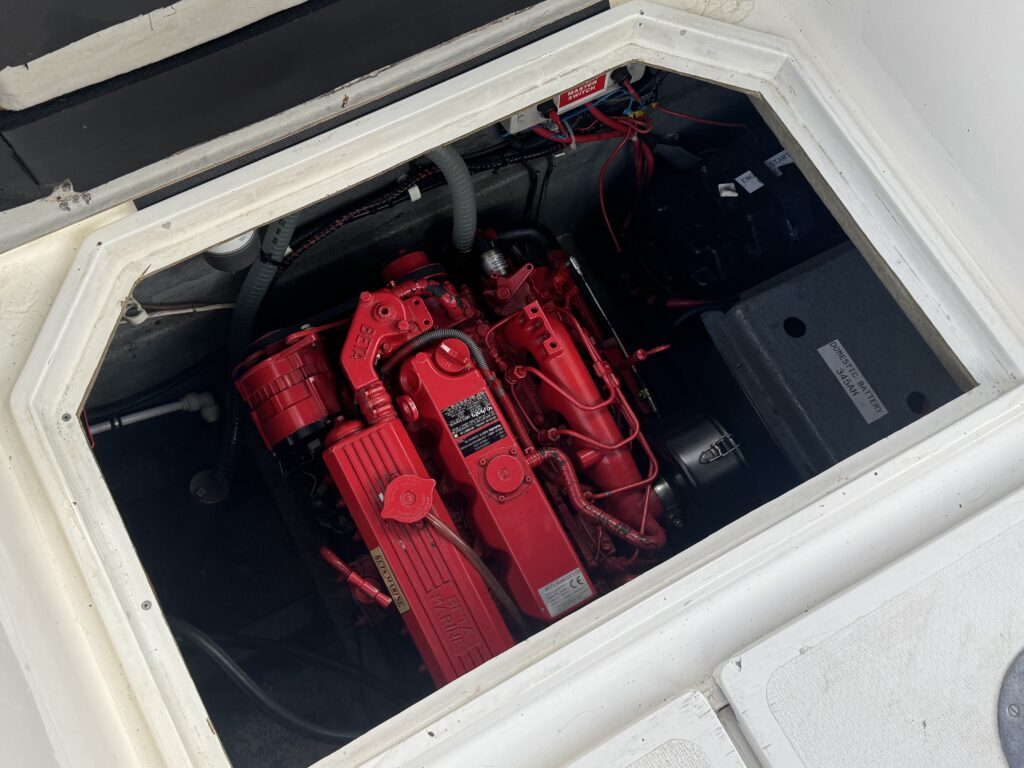 A close-up view of a boat engine compartment featuring a bright red engine with various hoses and wires. The engine is partially visible inside a rectangular opening with a white frame, showing the surrounding electrical components and battery.