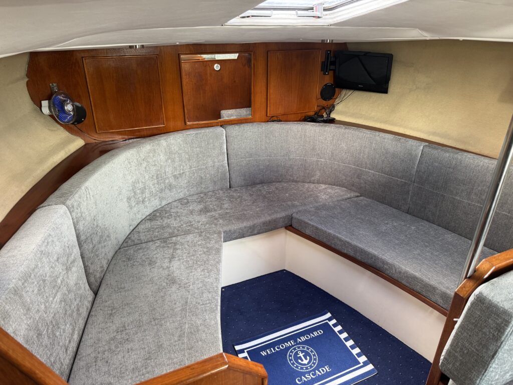 An interior view of a boat featuring a curved gray seating area with cushioned benches. A blue and white welcome mat is visible on the floor, and there is wooden cabinetry and a small television mounted on the wall.