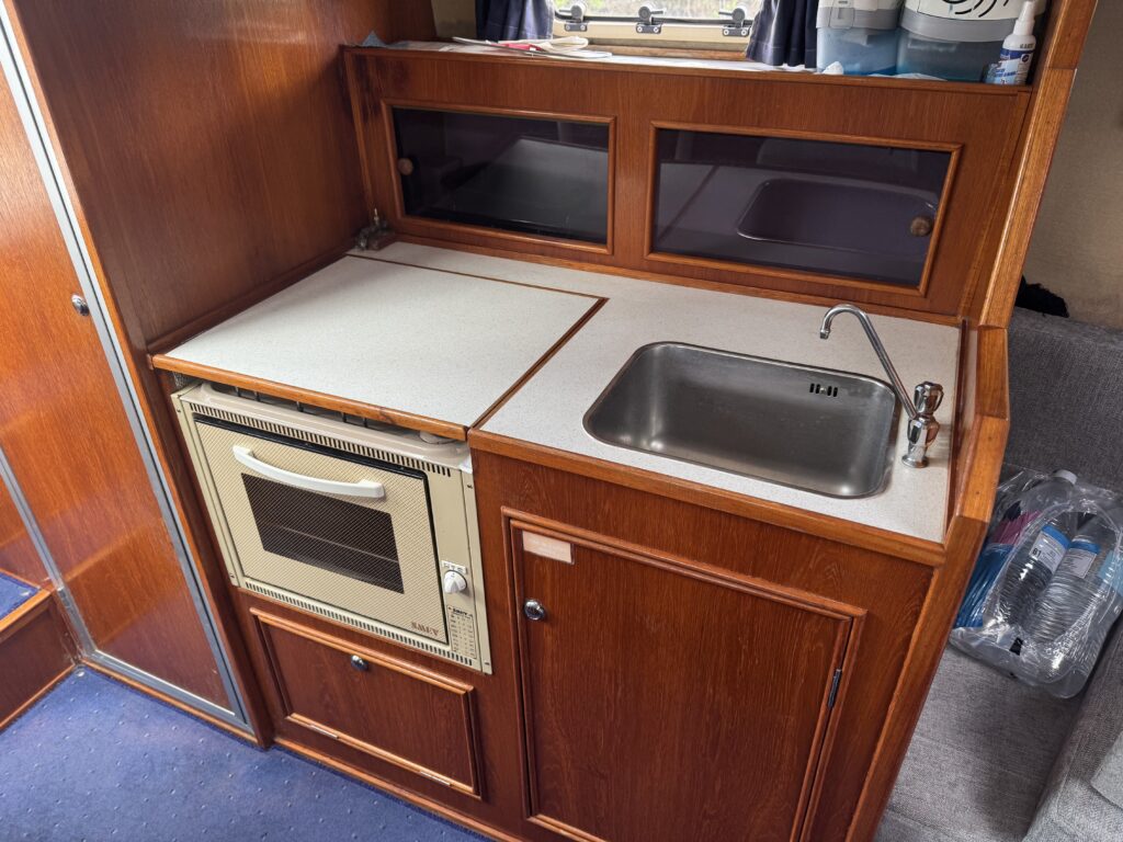 A compact kitchenette with wooden cabinetry, featuring a small oven below a countertop and a stainless steel sink beside it. The area has glass-fronted storage above the counter and is situated in a well-lit environment.