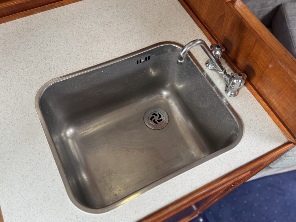 A stainless steel sink inset into a light-colored countertop, featuring a single faucet. The sink is rectangular with a drain at the bottom, and it is surrounded by wooden cabinetry. The view is from above, showing the sink's interior.