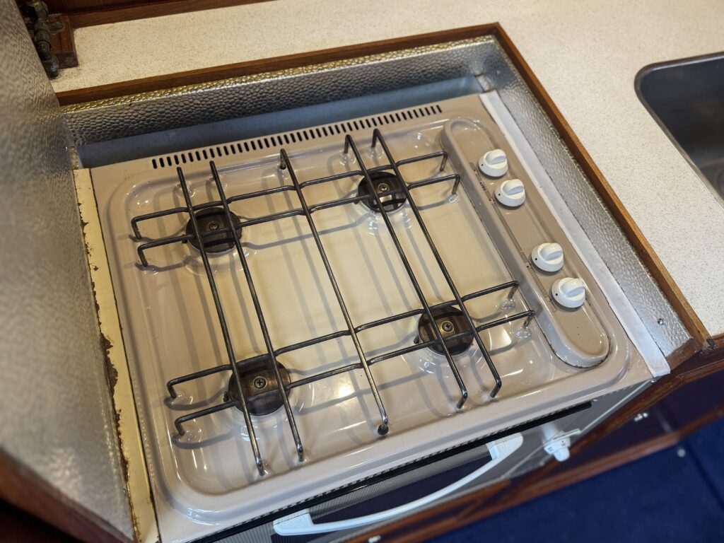 A compact gas stovetop with four burners set into a wooden kitchen countertop. The stove has a cream-colored surface with a metal grate and control knobs on one side, surrounded by a silver edge.