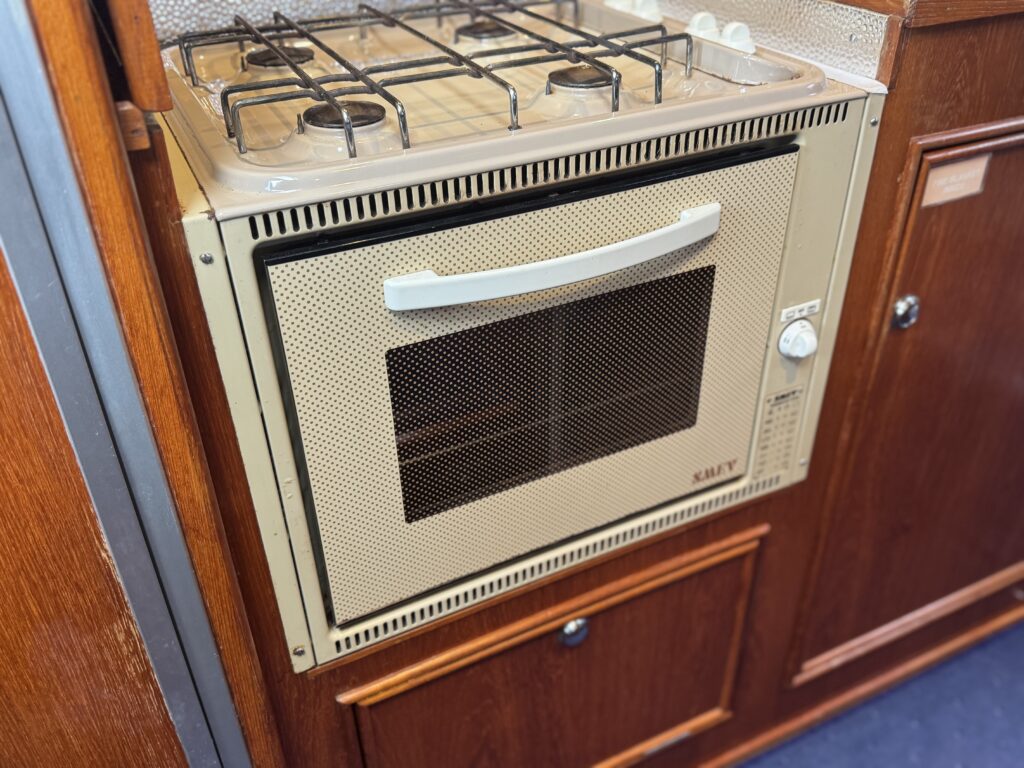 A beige gas oven with four burners on top and a mesh-covered window in front. It is installed in a wooden cabinet with a knob and an indicator light on the side. The surrounding cabinetry features a rich wood finish.
