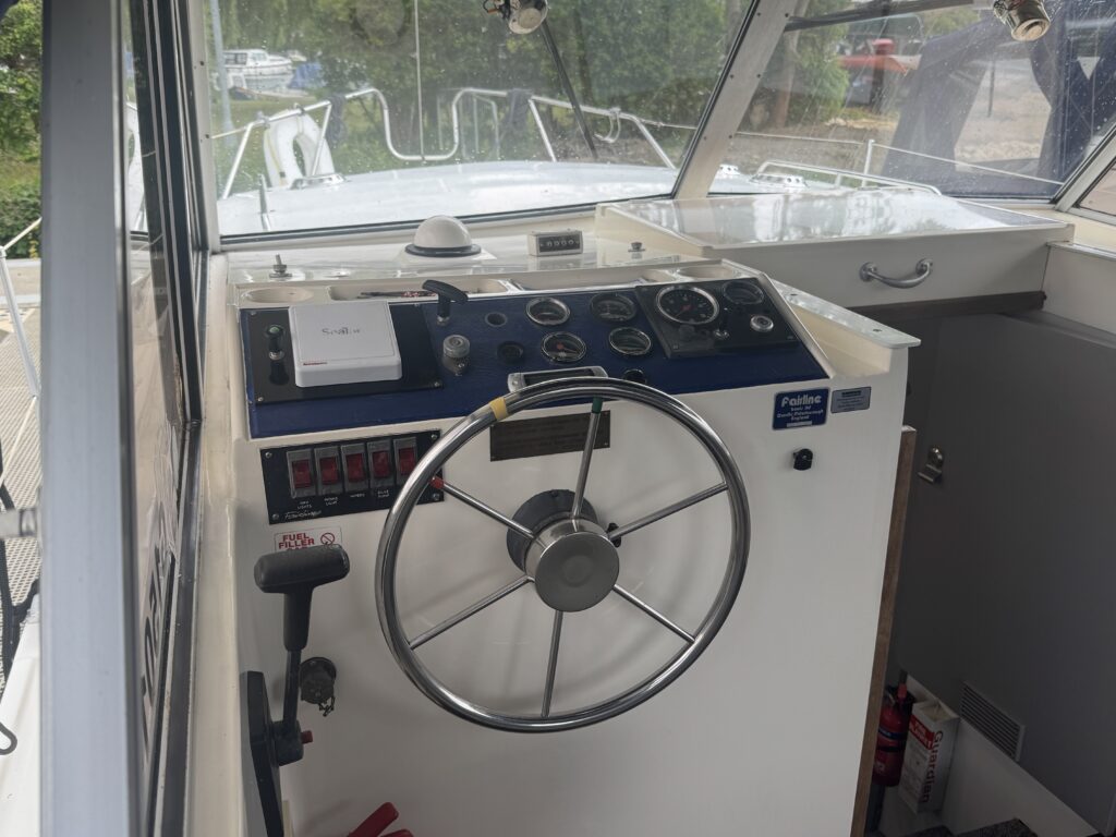 A boat's control panel featuring a steering wheel, various gauges, and switches mounted on a white dashboard. The scene shows a clear view through the front windows, with an outdoor area visible in the background.
