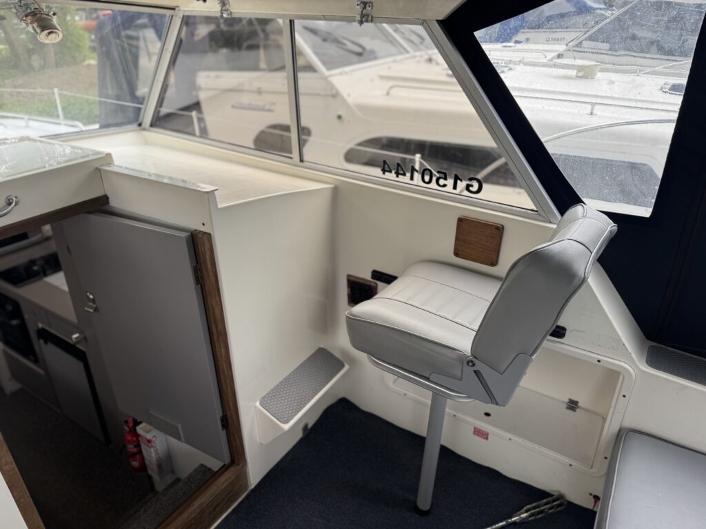 The interior of a boat cabin featuring a white swivel chair on a metal pedestal, a small counter area with storage underneath, and large windows that allow natural light. The floor is covered with dark carpet, and the background shows other boats docked outside.