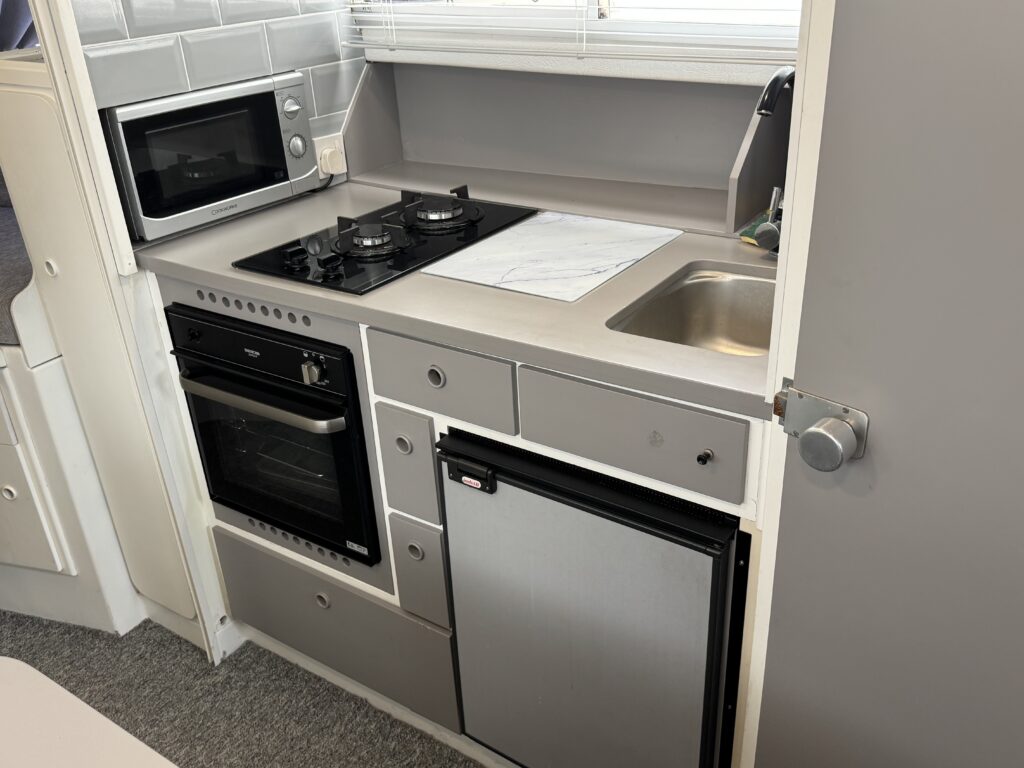 A compact kitchen featuring a microwave, a gas stove with two burners, a sink, and a small refrigerator. The cabinetry is light gray with a modern design, and the countertop is white with a marble pattern.