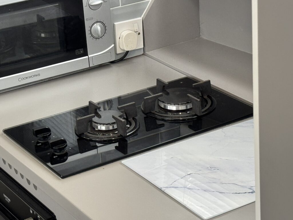 A modern kitchen with a black gas cooktop featuring two burners. The cooktop is placed on a light gray counter next to a white marble-patterned placemat. A gray microwave is visible in the background.