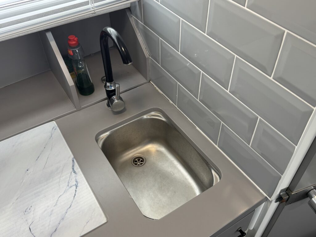 A stainless steel sink integrated into a light gray countertop, featuring a black faucet and a soap dispenser. The backsplash consists of glossy gray tiles, and there is a marble-patterned cutting board beside the sink.