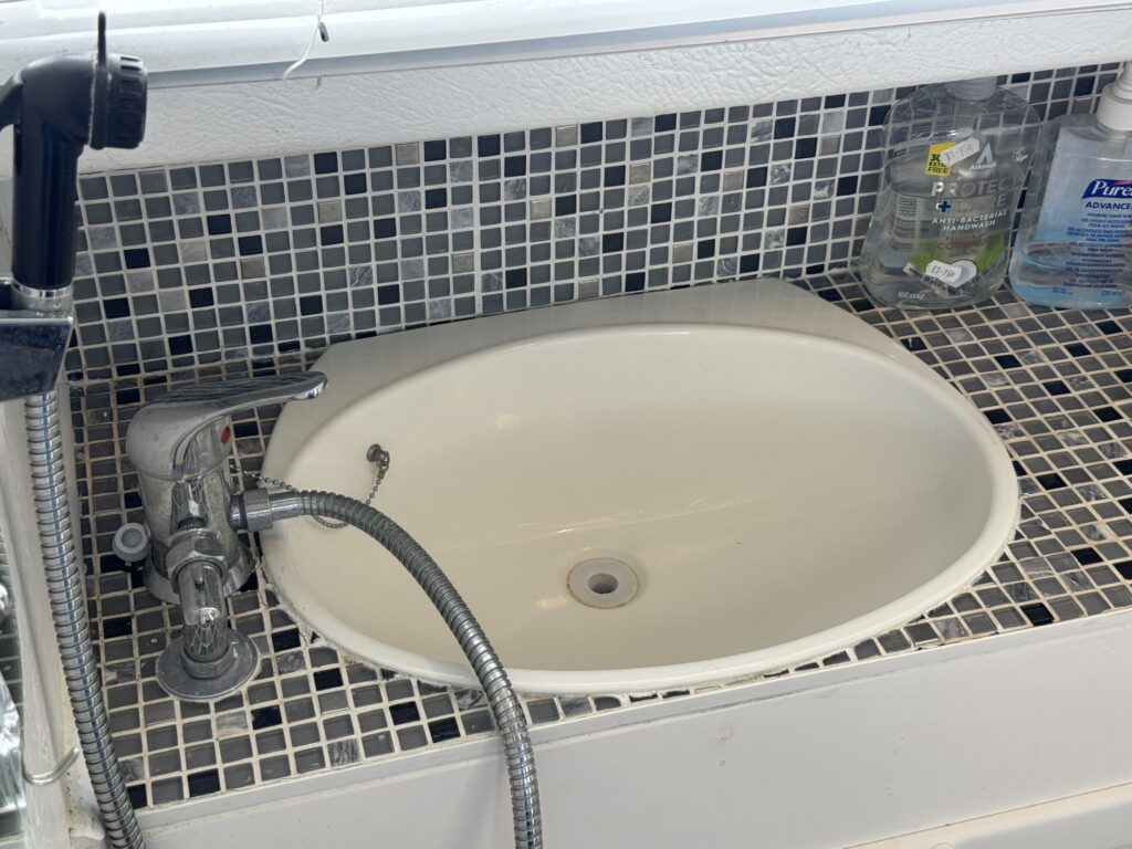 A white oval sink is mounted on a tiled surface made of small black and gray mosaic tiles. A faucet with a flexible hose is attached at the side, and bottles of liquid soap are visible in the background.