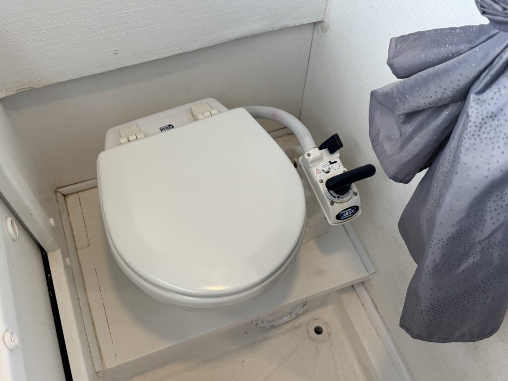 A compact white toilet with a closed lid positioned in a small bathroom corner. A control panel with levers is attached to the side of the toilet, and a curtain with droplets is partially visible in the background.