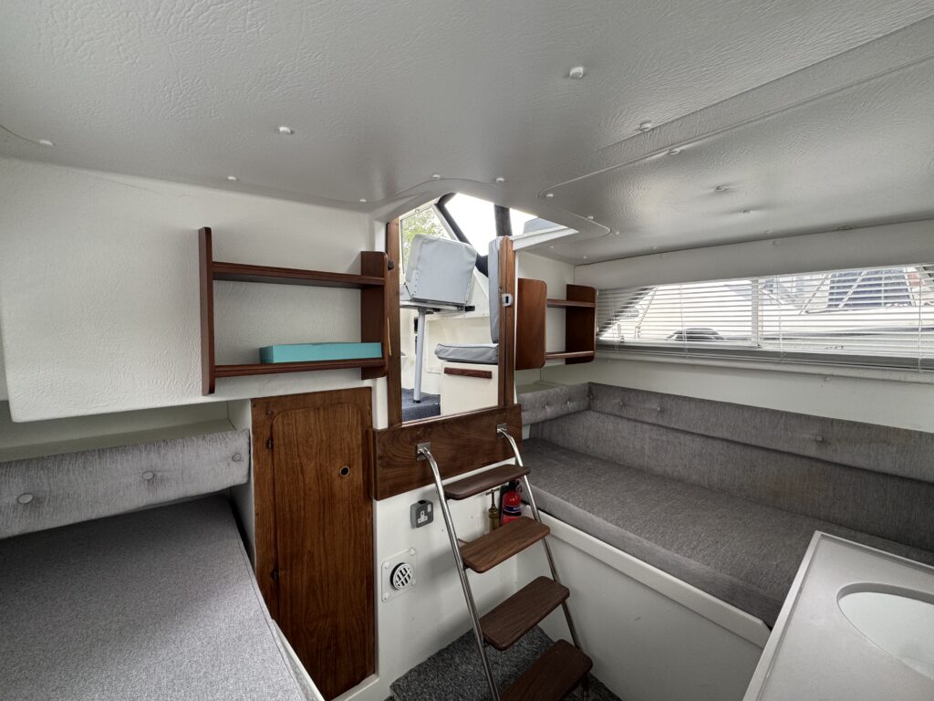 An interior view of a compact boat cabin featuring gray upholstered seating areas, wooden shelves, and a small ladder leading to an upper deck. Natural light filters in through windows, revealing storage spaces and a counter.