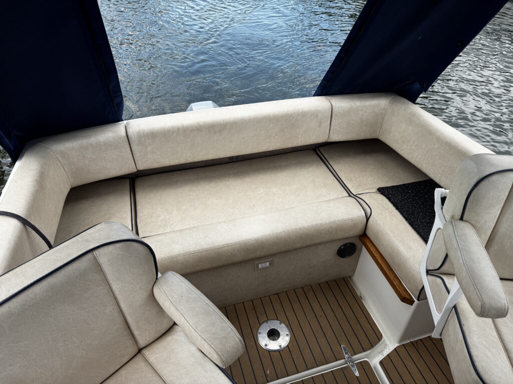 An overhead view of a light beige upholstered boat seating area with blue curtains. The seating wraps around the sides, and there is a textured mat on the floor near a circular drainage plate.