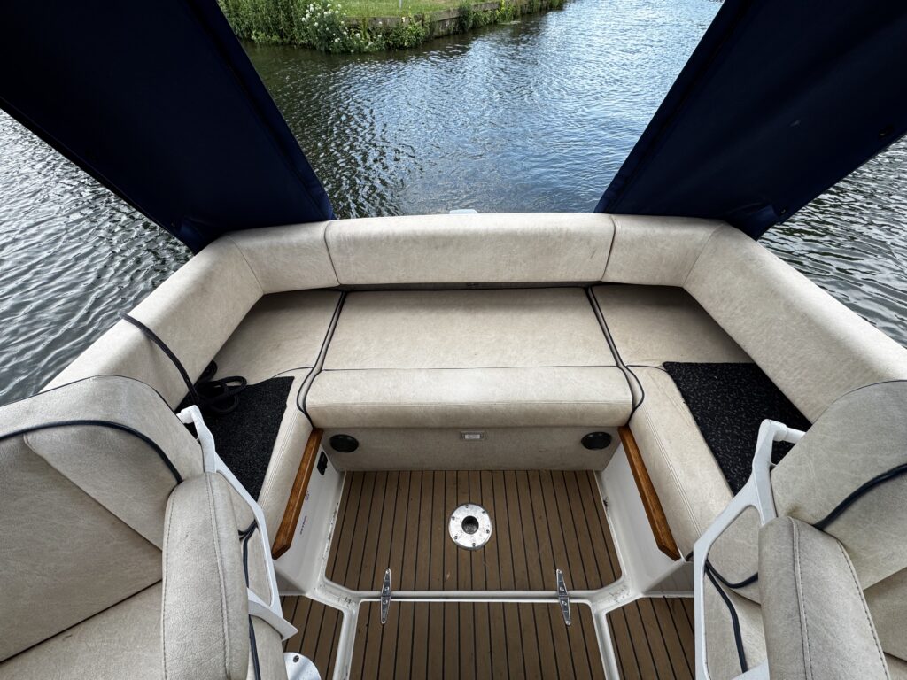 An overhead view of the interior of a boat featuring a beige seating area with cushions and a wooden floor. The scene includes two captain's chairs, and a view of calm water and greenery in the background.
