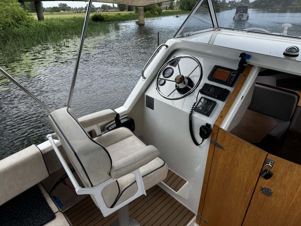The interior of a boat showing the helm, featuring a steering wheel, throttle controls, and navigation equipment. A cushioned captain's chair is positioned next to a wooden cabinet, with water and greenery visible outside the windows.