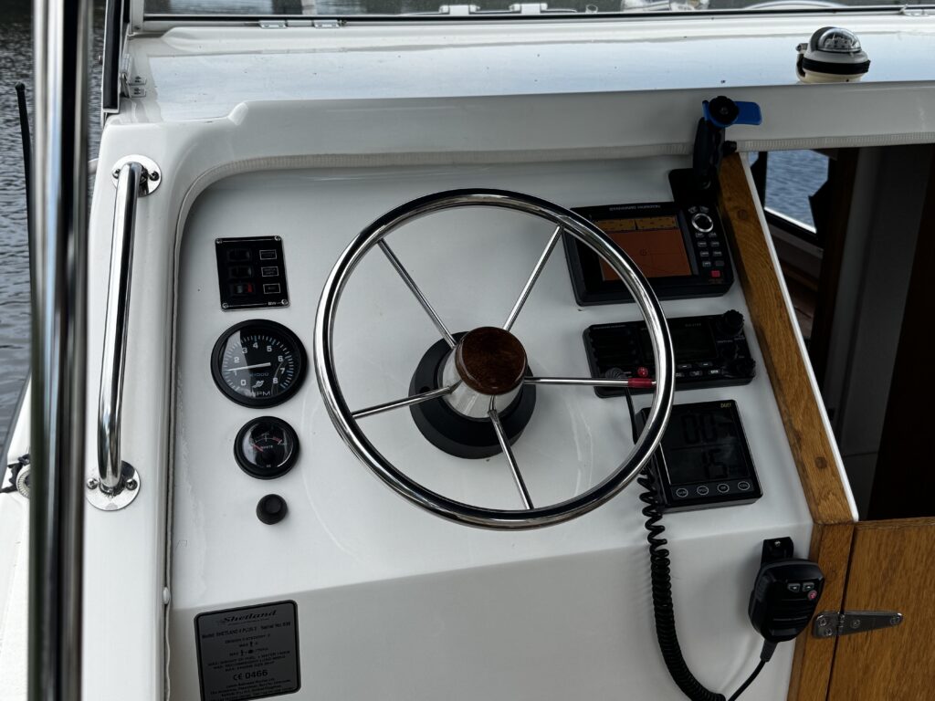 A close-up view of a boat's control panel featuring a steering wheel at the center. The panel includes various gauges, buttons, and a digital display, with a wooden section on the right. The background shows water and part of the boat's interior.