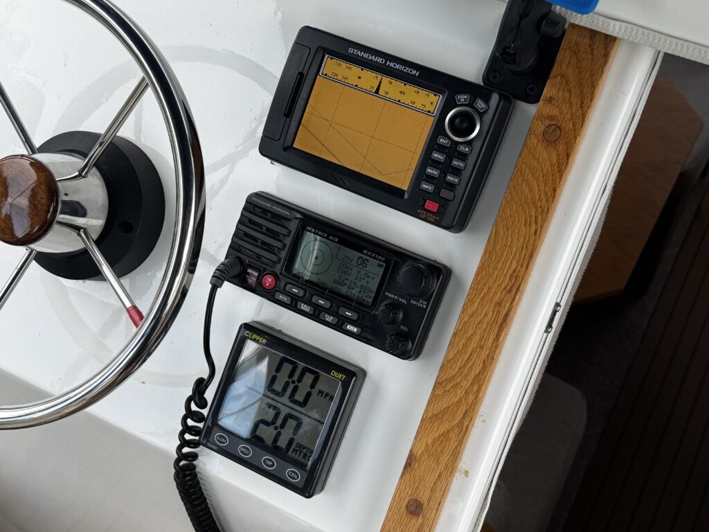 A marine control panel featuring three devices: a navigation display with a yellow screen at the top, a VHF radio in the middle with buttons and a display, and a digital thermometer beneath showing temperature and humidity readings. The boat's steering wheel is visible to the left.