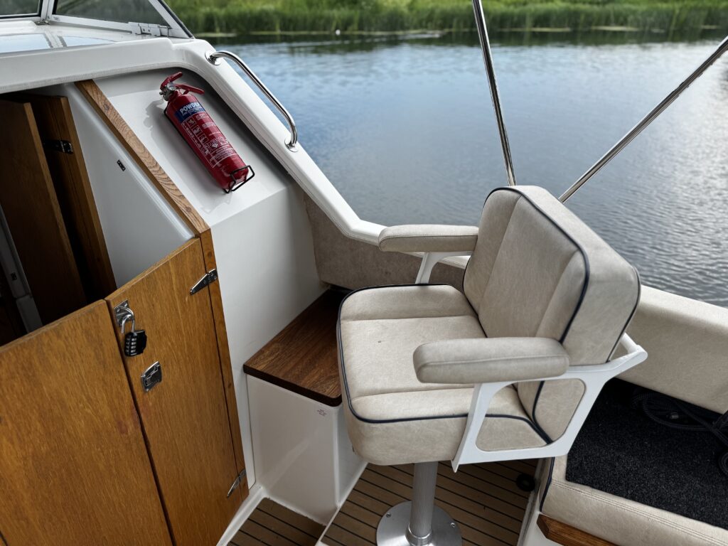 A light-colored padded captain's chair is positioned next to a wooden cabinet on a boat. A fire extinguisher is mounted on the wall above the chair. The background shows calm water and green vegetation.