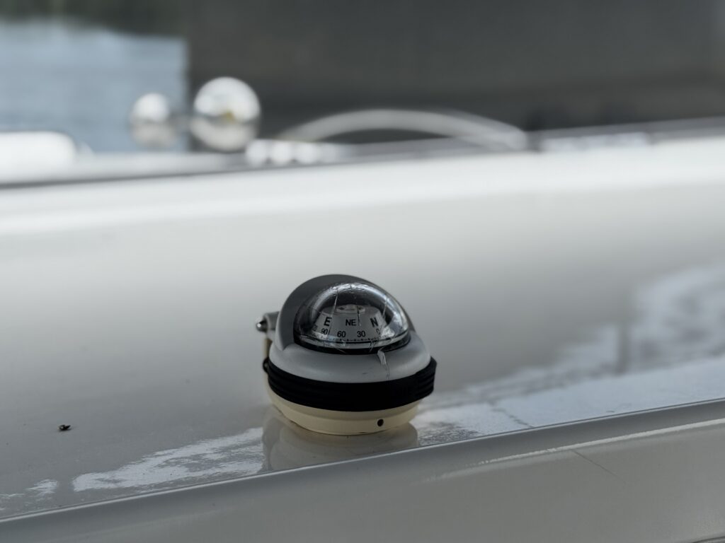 A small compass mounted on a white surface, featuring a clear, domed cover and a black base. The compass needle is visible, indicating direction, while the background shows a blurred view of water and reflections.